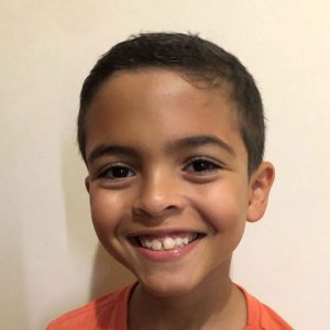 Samuel é um menino branco, de cabelos escuros e curtos. Veste camiseta laranja. Ao fundo, parede branca