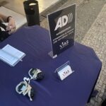 Placa de acrílico informando do serviço de AD, ao lado de aparelhos de audiodescrição e cartões de visita da Vias Acessíveis, sobre uma mesa com toalha azul