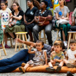 3 crianças sentadas no chão e alguns adultos em cadeiras, ao fundo