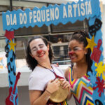 Duas palhaças sorriem abaixo de um pórtico azul escrito "dia do pequeno artista"