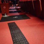 vista interna da sala de cinema. Sobre piso carpetado vermelho, pisos táteis pretos e demarcação para cadeira de rodas