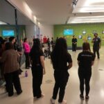 turma de pé, em roda. Ao centro, instrutor sinaliza em libras. Estão em um saguão espaçoso.