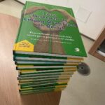 pilha de livros com título "Livro Ilustrado da Língua Brasileira de Sinais". A capa é verde com mãos formando um coração.