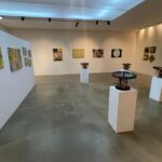 quadros em paredes brancas e quatro obras táteis ao centro, em uma sala ampla de exposição
