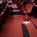 vista interna de uma das fileiras da sala de cinema. Sobre piso carpetado vermelho, pisos táteis pretos e duas demarcações para cadeira de rodas