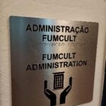 placa em chapa de metal com fontes pretas e braille da Administração FUMCULT