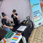 Duas mulheres da equipe atrás de uma mesa com equipamentos de audiodecrição e outros objetos ligados ao universo braille