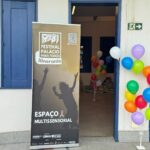 banner do Festival Palácio para Todos na cor marrom com dizeres em branco, ao lado de uma porta. Do outro lado, suporte com bexigas coloridas