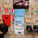 Banner Inclua em tom de azul com dizeres coloridos, fixado em parede de azulejos de vários padrões e cores.