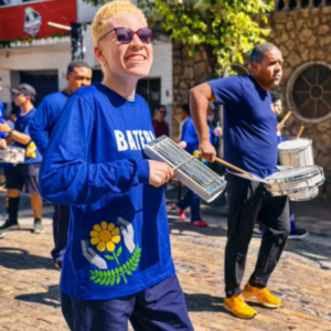 pessoa branca de cbelos loiros curtos e óculos escuros toca reco-reco na bateria da Apae Caratinga, durante desfile de rua