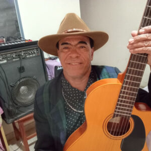 Carlos Brumm é homem pardo e sobrancelhas grossas. Segura viola na frente do troncho e veste chapéu de vaqueiro marrom.