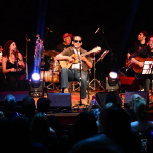 Danilo Bayão toca violão no palco com banda. Ele usa óculos escuros e está sentado num banquinho.