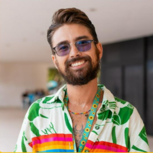 Peu Avelar é um jovem branco de cabelos castanhos claros e curtos. Usa óculos escuros e tem barba. Veste camisa com estampas coloridas e o cordão de girassol.
