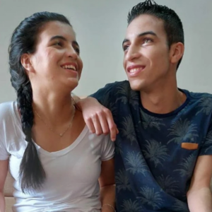 Leandro e Sandy sorriem lado a lado. Ambos são jovens, têm pele clara, cabelos pretos e têm deficiência visual.