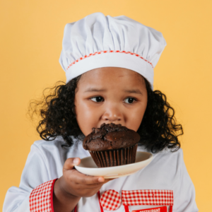 Foto de criança parda e cabelos cacheados veste uniforme de chef. Ela segura um prato com muffin de chocolate
